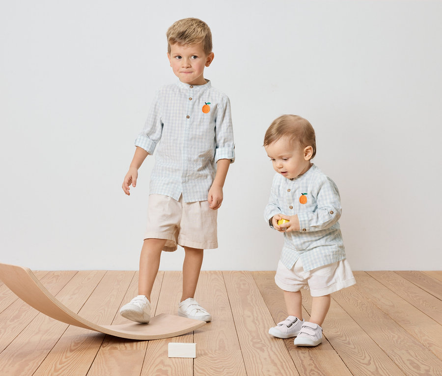 Deux enfants, un garçon plus âgé debout sur une bascule en bois et un petit garçon tenant une balle jaune, tous deux portant des chemises à carreaux avec une application orange.