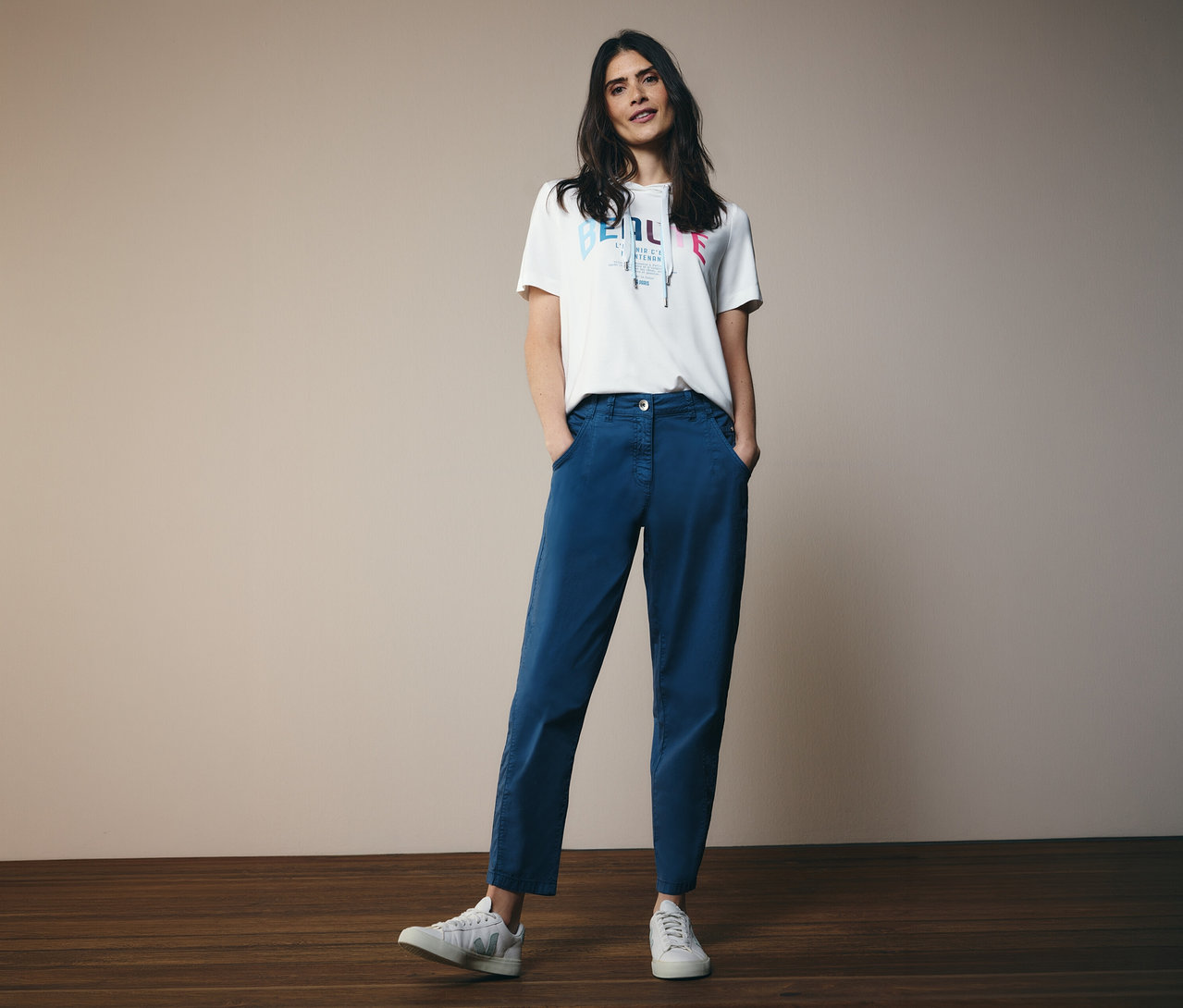 Une femme portant un t-shirt à capuche CECIL et un barrel CECIL « Scarlett » bleu, les mains dans les poches, pose devant la caméra.