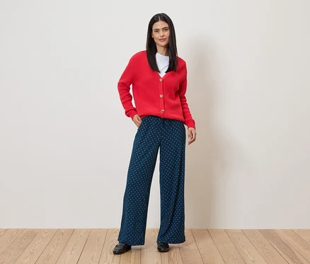 Une femme portant un cardigan rouge et un pantalon bleu marine à pois blancs se tient sur un plancher en bois devant un mur.