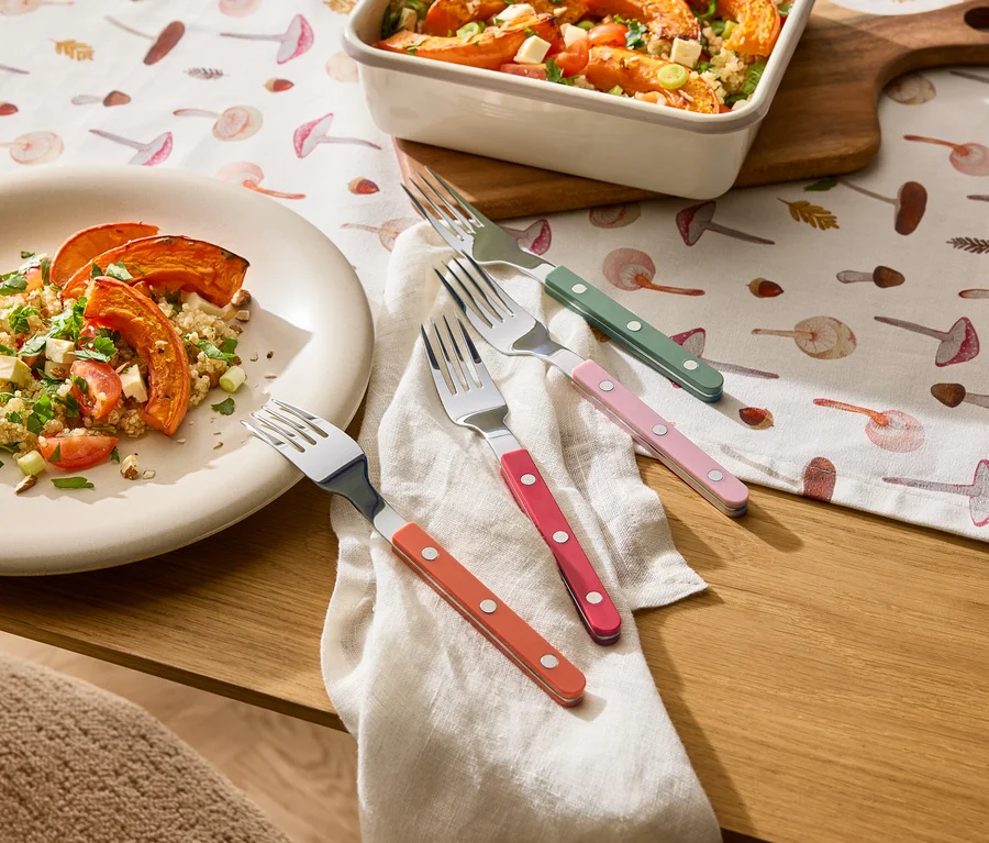 Une assiette et un plat à gratin avec un plat à base de courge sont posés sur une nappe à motifs de champignons, à côté de quatre fourchettes à manches colorés sur un tissu.