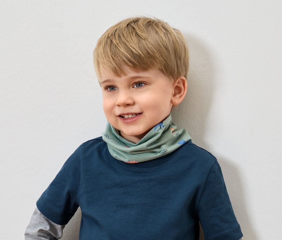 Un garçon souriant aux cheveux blonds et portant un t-shirt bleu porte un foulard vert à motifs animaliers.