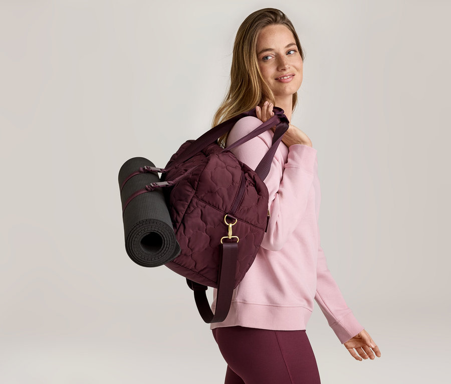 Femme avec un sac de sport et un tapis de yoga sur l'épaule, portant un pull rose et un legging bordeaux.