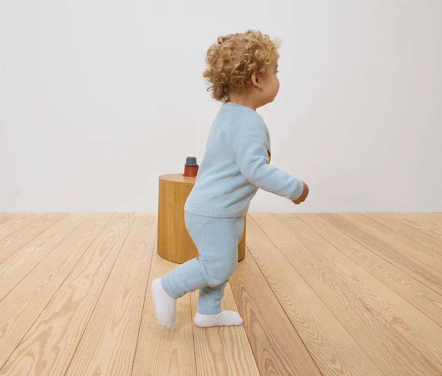 Un jeune enfant aux cheveux bouclés marche sur un plancher en bois. Il porte un pull bleu clair et un pantalon assorti.
