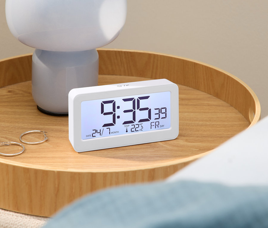 Réveil numérique affichant 9h35 sur une table en bois avec une lampe et des boucles d'oreilles.