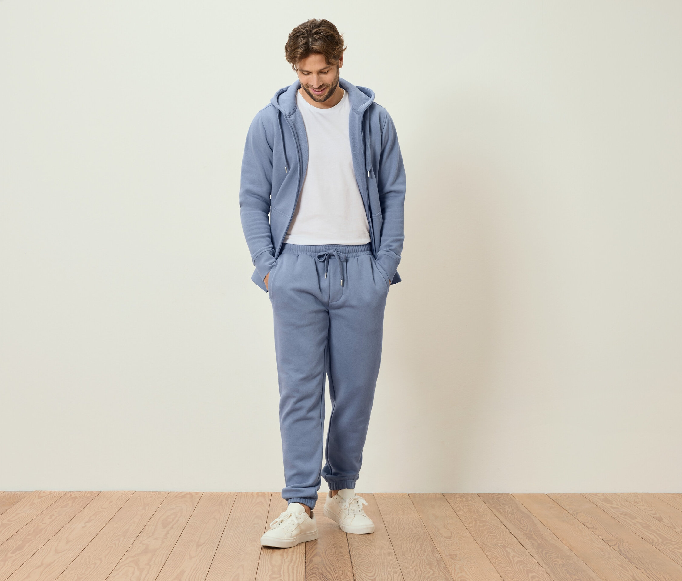 Homme debout, les mains dans les poches, en survêtement bleu clair et baskets blanches.