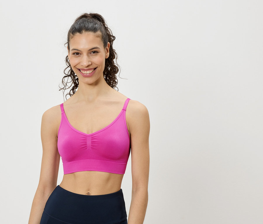 Une femme souriante aux cheveux bouclés relevés porte un soutien-gorge de sport rose et un legging bleu marine sur un fond blanc.