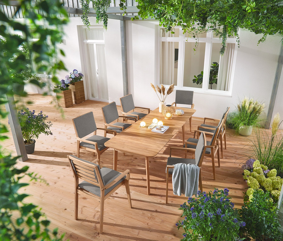 Une table à manger extensible en teck avec huit chaises de jardin empilables en teck se trouve sur une terrasse, décorée de deux colonnes à plantes Tinus, d'une lanterne solaire en bois ronde et de trois piquets solaires Hortensie.