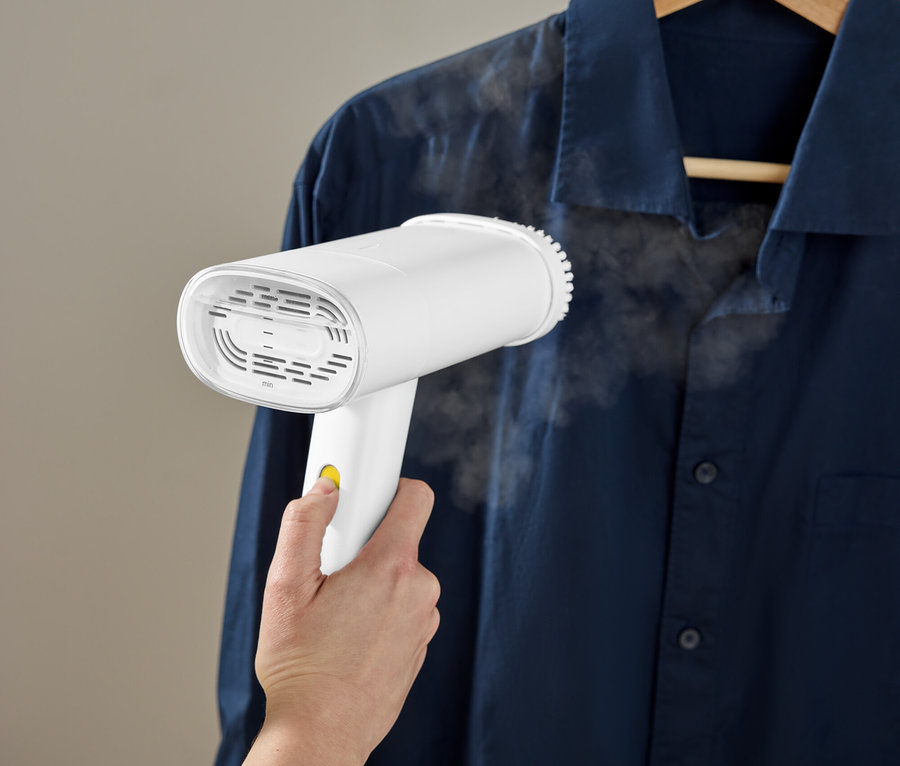 Une main utilise un défroisseur blanc pour lisser une chemise bleue suspendue à un cintre en bois.