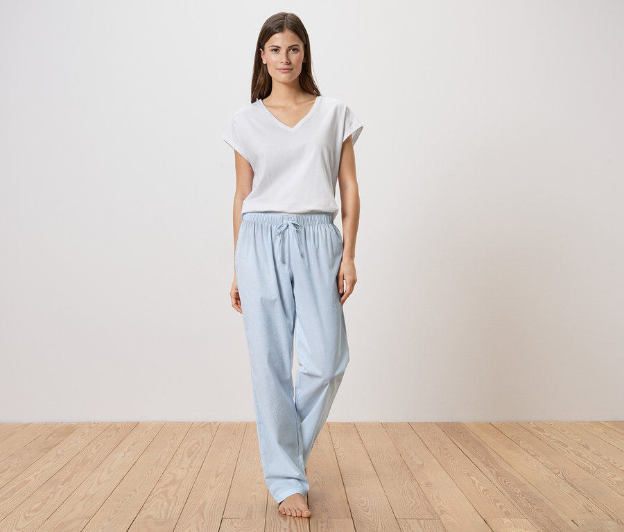 Une femme se tient pieds nus sur un plancher en bois, portant un t-shirt blanc et un pantalon de pyjama bleu clair.