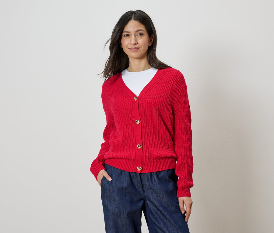 Femme portant un cardigan rouge avec des boutons en forme de cœur, un t-shirt blanc et un pantalon bleu.