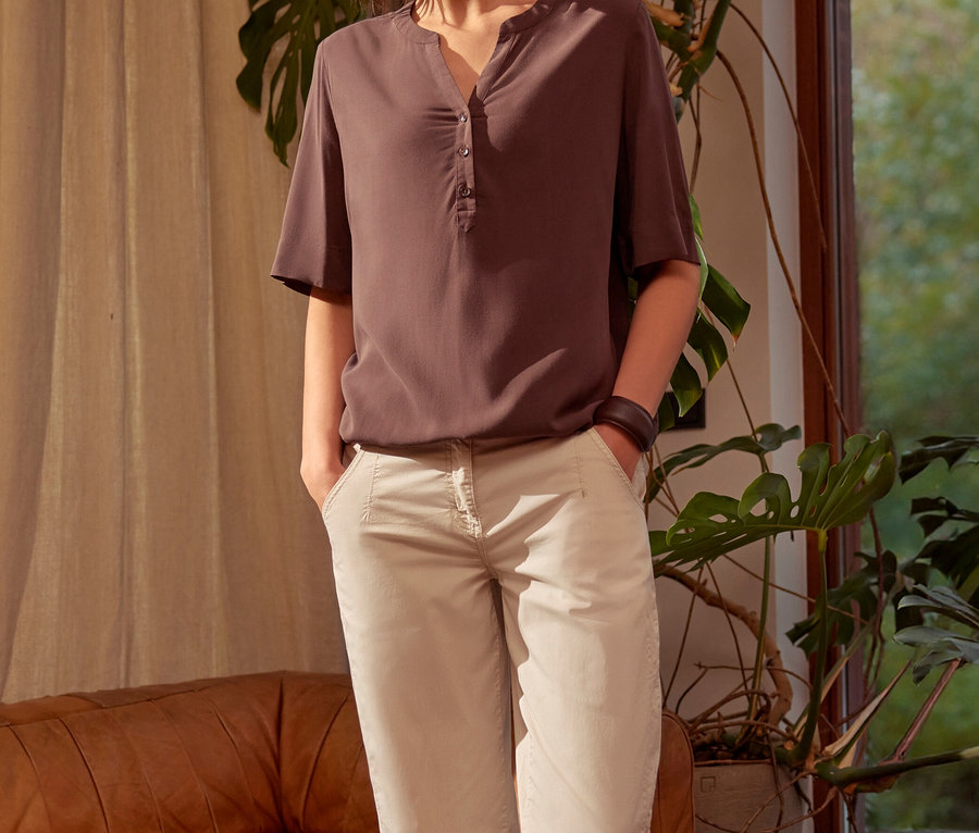 Femme debout les mains dans les poches, portant un chemisier tunique CECIL marron et un pantalon clair, dans un salon avec un canapé en cuir et une plante.