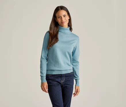 Une femme aux longs cheveux bruns porte un pull à col roulé bleu clair et un jean foncé sur un fond clair.