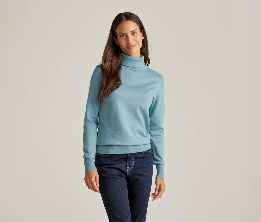 Une femme aux longs cheveux bruns porte un pull à col roulé bleu clair et un jean foncé sur un fond clair.