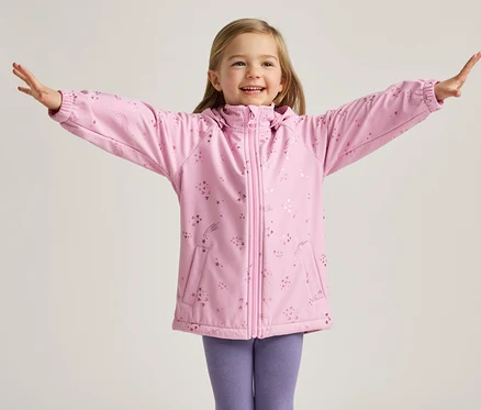 Une fillette blonde souriante se tient les bras écartés, vêtue d'une veste rose à motifs d'étoiles et d'un legging violet, sur un fond clair.