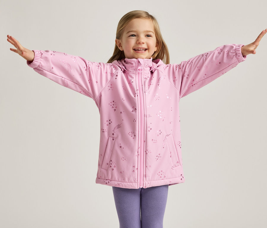 Une fillette blonde souriante se tient les bras écartés, vêtue d'une veste rose à motifs d'étoiles et d'un legging violet, sur un fond clair.