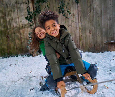 Deux enfants souriants sur une luge en bois dans la neige. Un garçon en doudoune verte est assis devant, tenant la corde de la luge, avec une fille en haut vert assise derrière lui. Ils sont devant une clôture en bois.