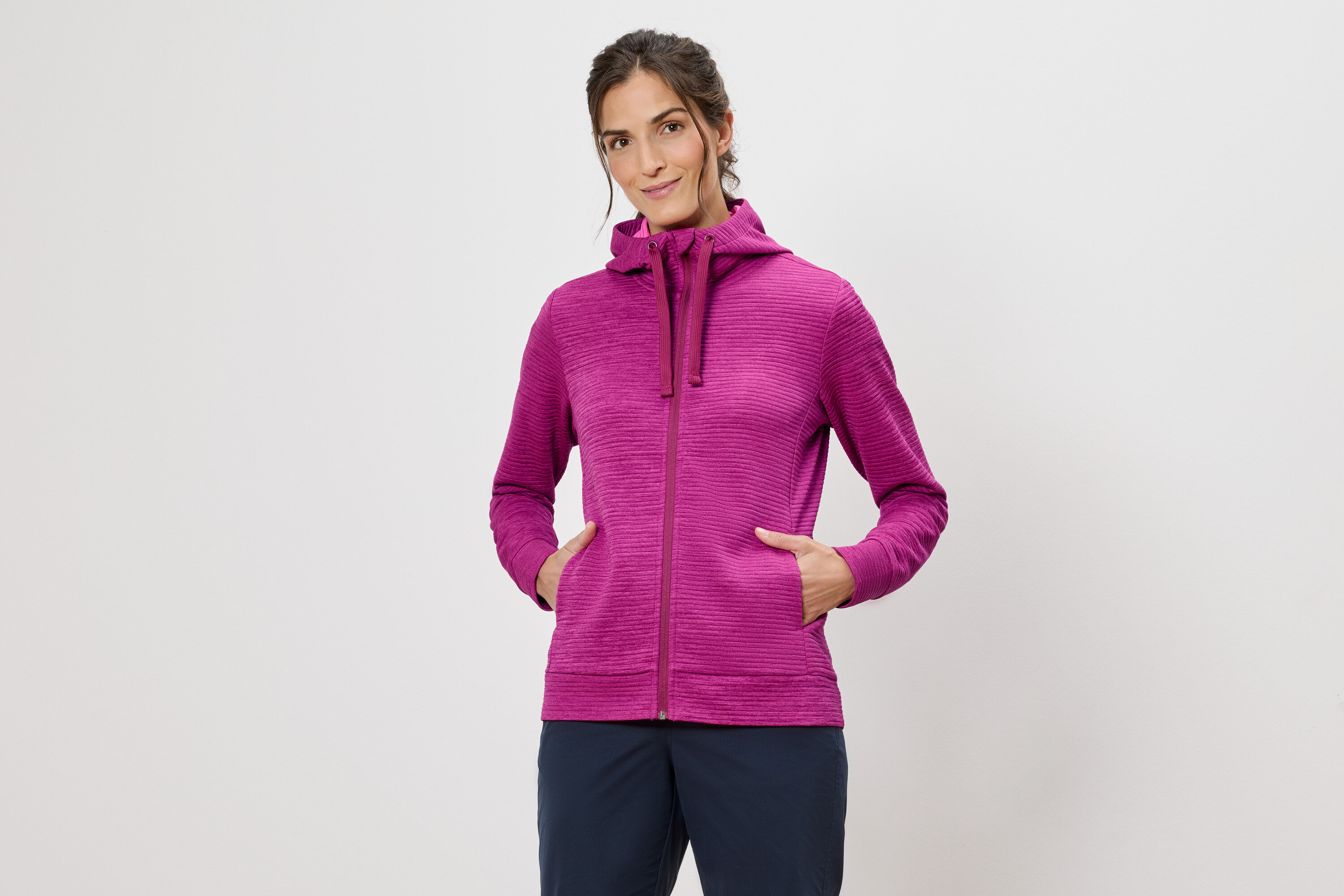 Femme portant un sweat à capuche rose, les mains dans les poches, sur un fond blanc.