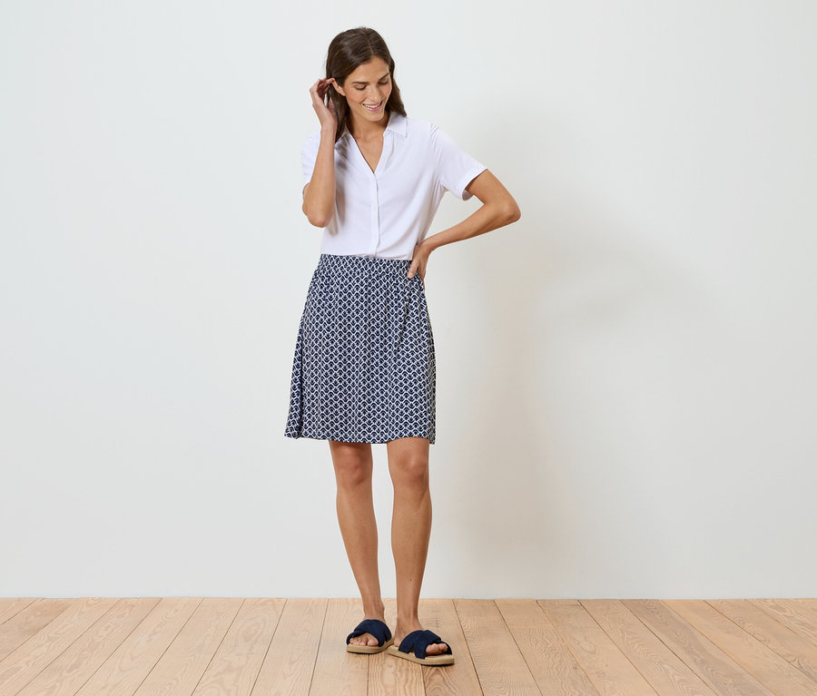 Femme debout devant un mur blanc portant une chemise blanche et une jupe à motifs bleu et blanc.