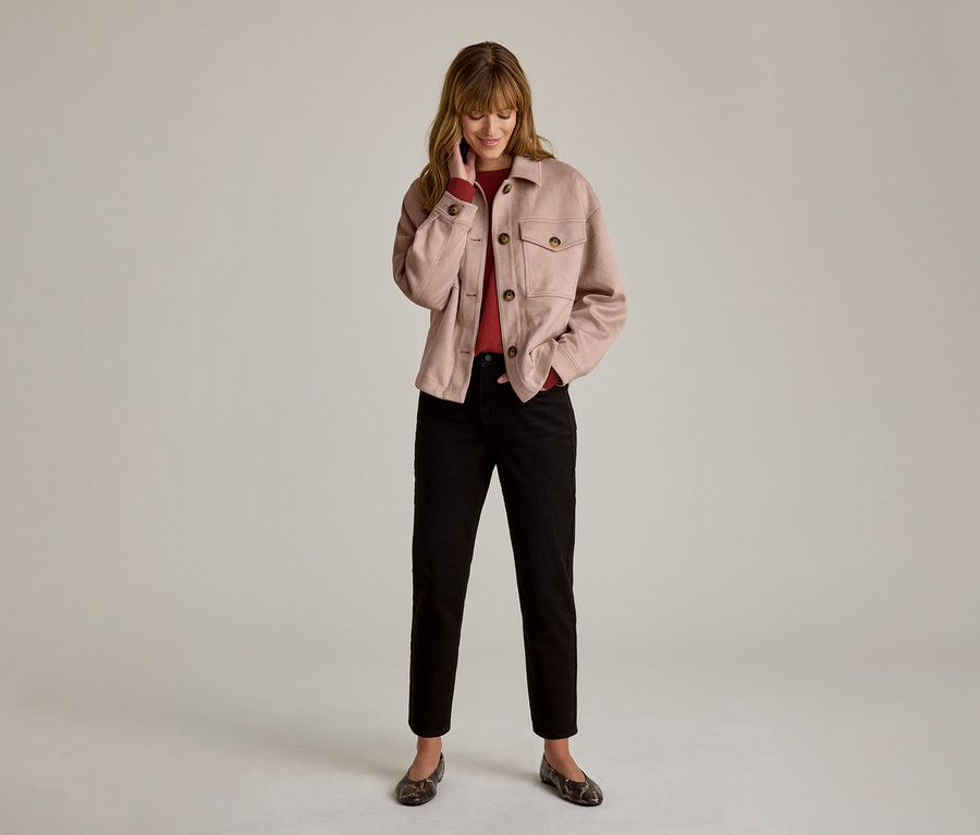Femme en veste rose clair, haut rouge, pantalon noir et ballerines à motifs, souriant et regardant vers le bas, une main près du menton.