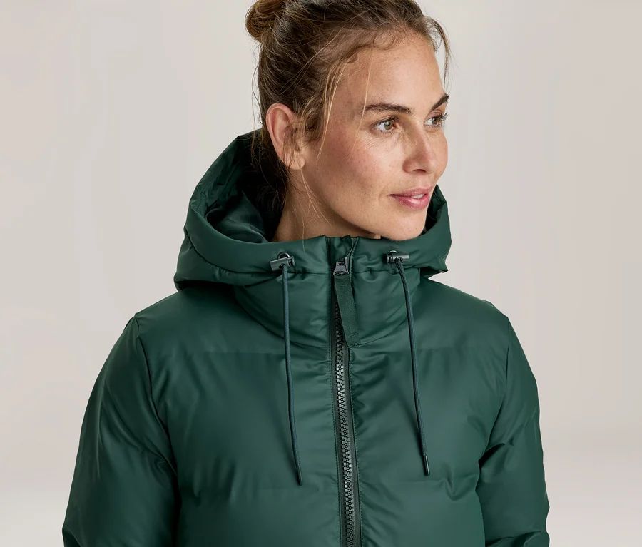Femme portant une doudoune vert foncé avec capuche, regardant vers la droite. Ses cheveux sont attachés en chignon.
