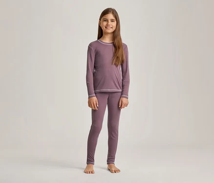 Une fille souriante aux longs cheveux bruns est debout pieds nus, vêtue d'un pyjama mauve à manches longues et d'un legging assorti, avec des coutures décoratives gris clair.