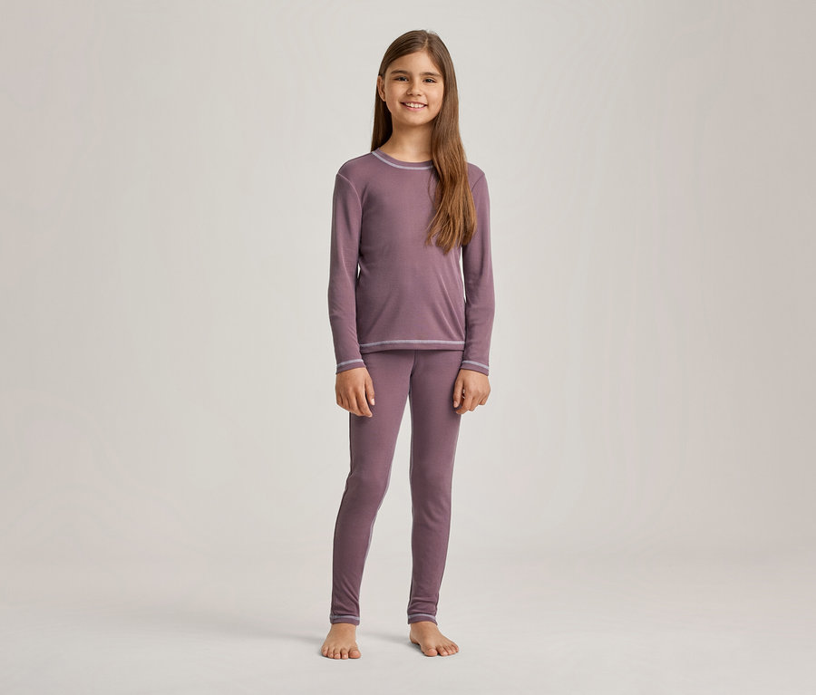 Une fille souriante aux longs cheveux bruns est debout pieds nus, vêtue d'un pyjama mauve à manches longues et d'un legging assorti, avec des coutures décoratives gris clair.