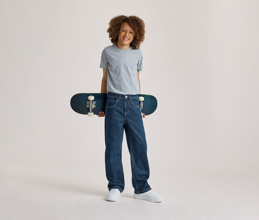 Garçon souriant aux cheveux bouclés, portant un t-shirt gris, un jean bleu et des baskets blanches, tenant un skateboard bleu foncé derrière son dos.