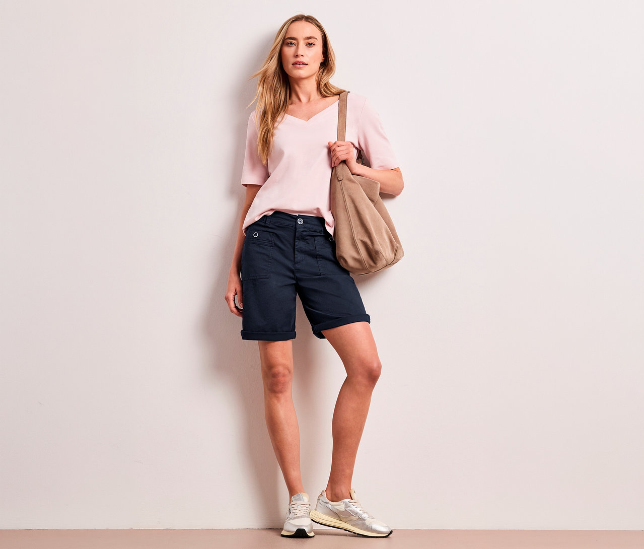 Une femme aux cheveux blonds porte un haut rose pâle, un bermuda Street One « Yulius » bleu marine et des baskets claires, tenant un sac à bandoulière beige.