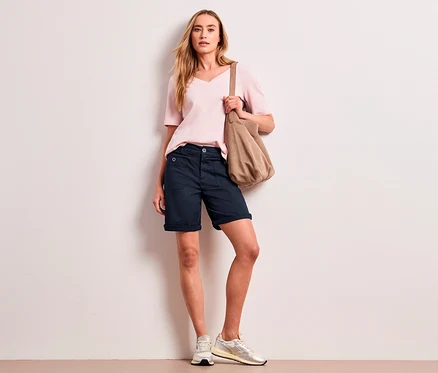 Une femme aux cheveux blonds porte un haut rose pâle, un bermuda Street One « Yulius » bleu marine et des baskets claires, tenant un sac à bandoulière beige.