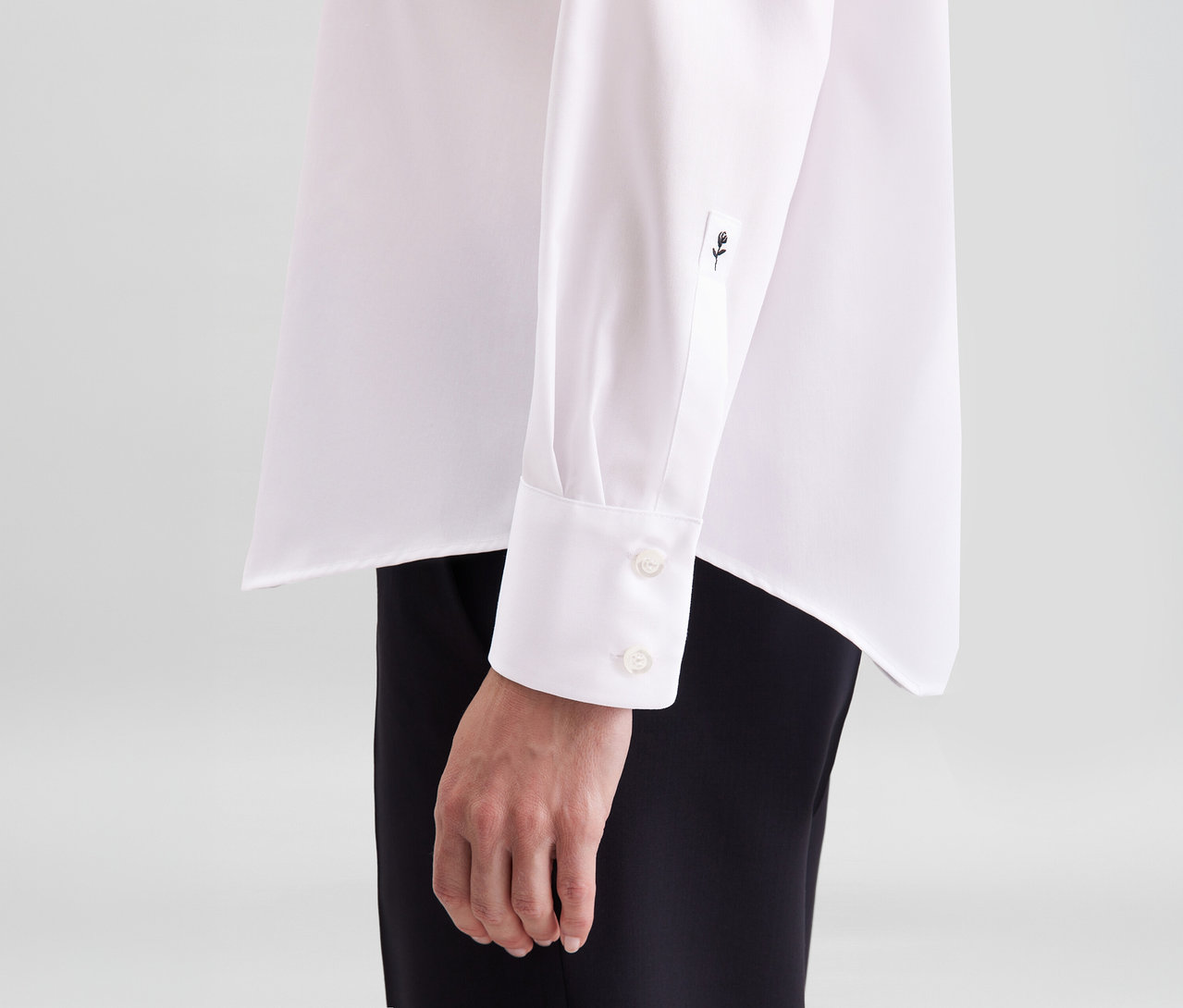 Gros plan d'une personne portant un chemisier sans repassage Seidensticker blanc avec un poignet à deux boutons et un petit logo rose brodé sur la manche, associé à un pantalon noir.