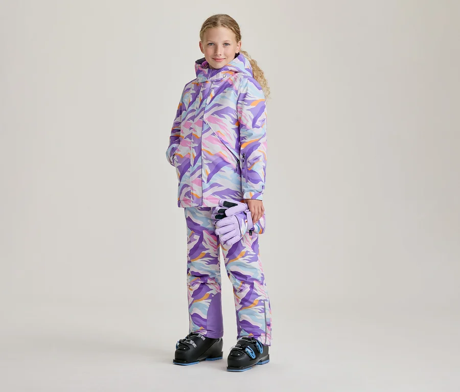 Une jeune fille aux cheveux blonds et bouclés sourit, vêtue d'une veste de ski colorée et d'un pantalon de ski assorti avec un motif abstrait violet, rose, bleu et orange. Elle tient des gants violet clair dans sa main gauche et porte des chaussures de ski noires avec des accents bleus.