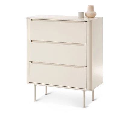 Une commode beige clair à trois tiroirs, chacun avec une poignée encastrée. Elle a des coins arrondis et repose sur quatre pieds fins. Un petit objet cylindrique gris et un vase beige bulbeux sont posés dessus.