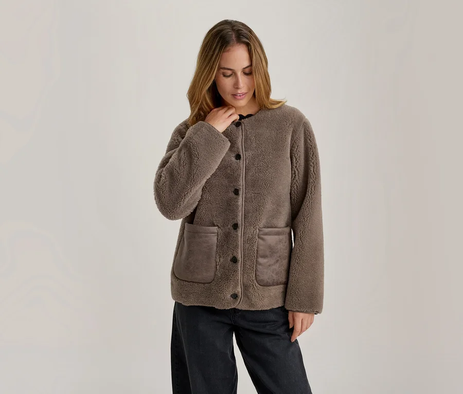 Femme portant une veste teddy marron avec des boutons foncés et deux poches avant, associée à un pantalon foncé, regardant vers le bas et ajustant son col.