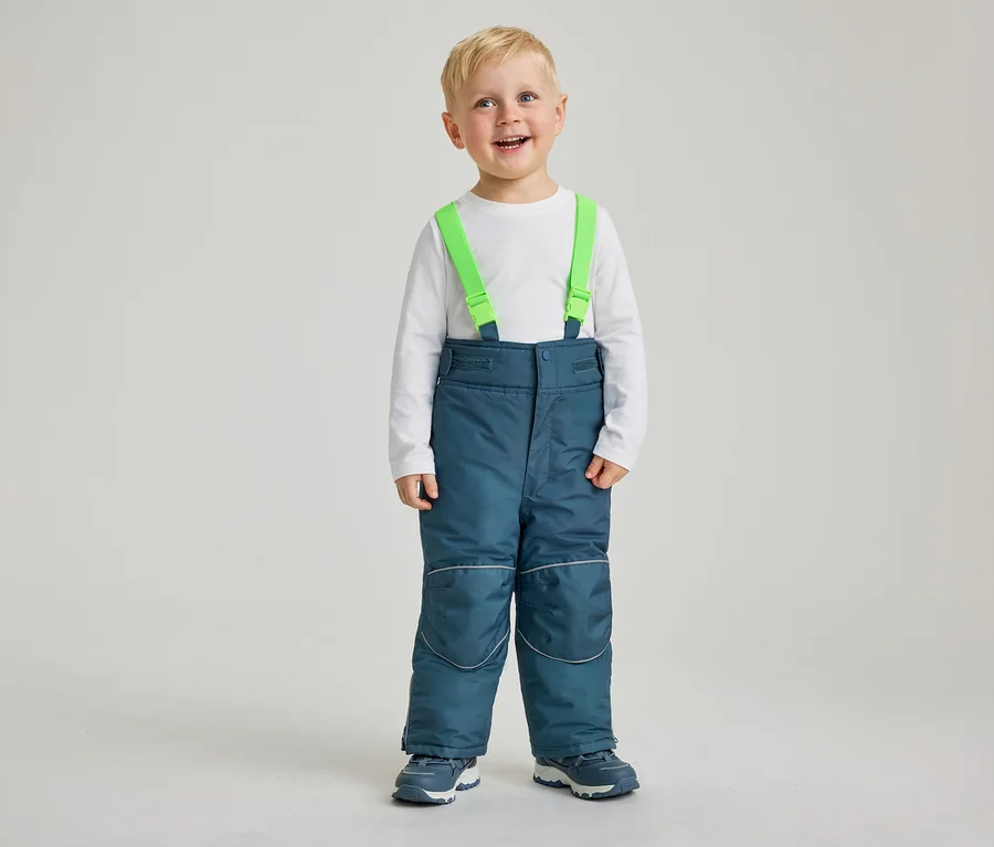 Un jeune garçon aux cheveux blonds sourit à la caméra, vêtu d'un t-shirt blanc à manches longues, d'un pantalon de neige bleu avec des bretelles vertes et de baskets bleues et blanches. Le pantalon de neige a des bandes réfléchissantes aux genoux et aux chevilles.