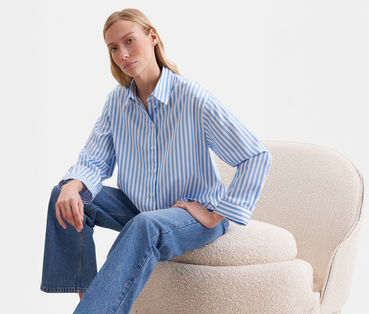 Femme blonde assise sur un fauteuil beige, portant un chemisier oversize Seidensticker rayé bleu et blanc et un jean bleu.