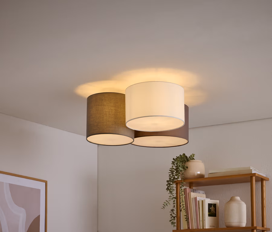 Trois plafonniers avec des abat-jour cylindriques blancs, gris foncé et marron sont regroupés sur un plafond blanc. Une étagère en bois avec des livres et une plante est visible en bas à droite, et un tableau abstrait encadré en bas à gauche.