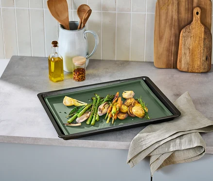 Des légumes comme des asperges, des carottes et des pommes de terre sont posés sur une plaque de cuisson verte. À côté se trouvent de l'huile, des épices et des ustensiles de cuisine.