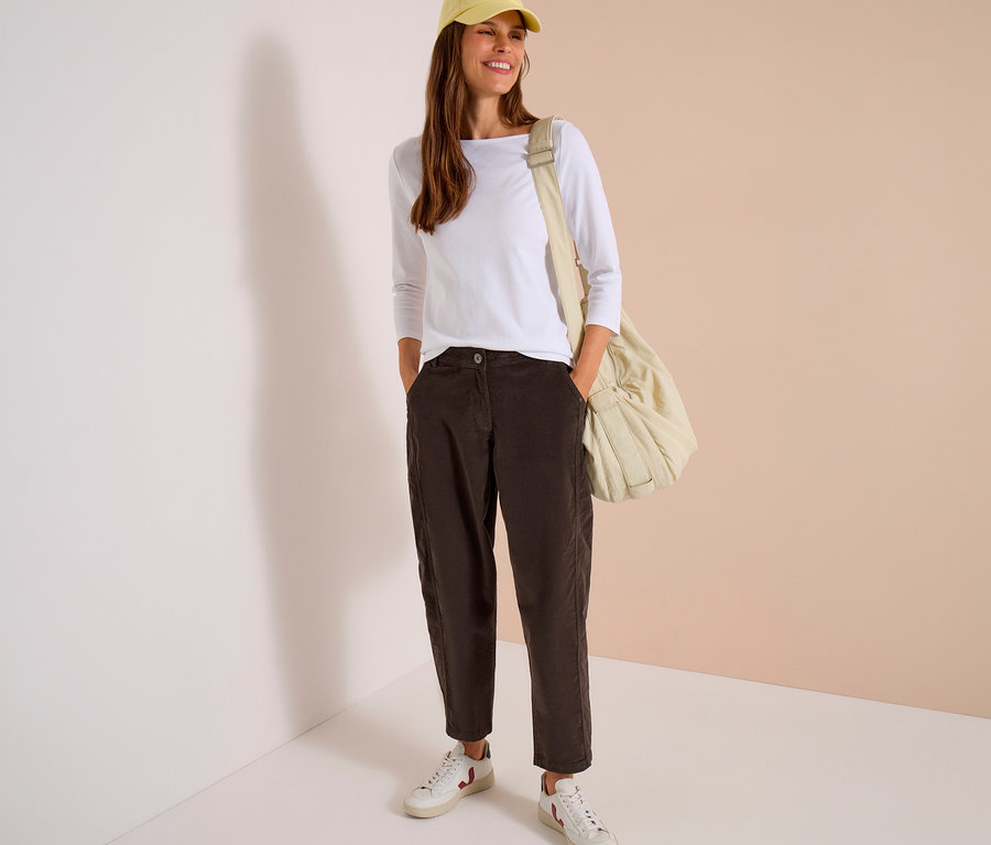 Femme souriante portant une casquette jaune, un haut blanc à manches longues, un pantalon en velours côtelé CECIL marron foncé, mains dans les poches, des baskets blanches et un sac beige.