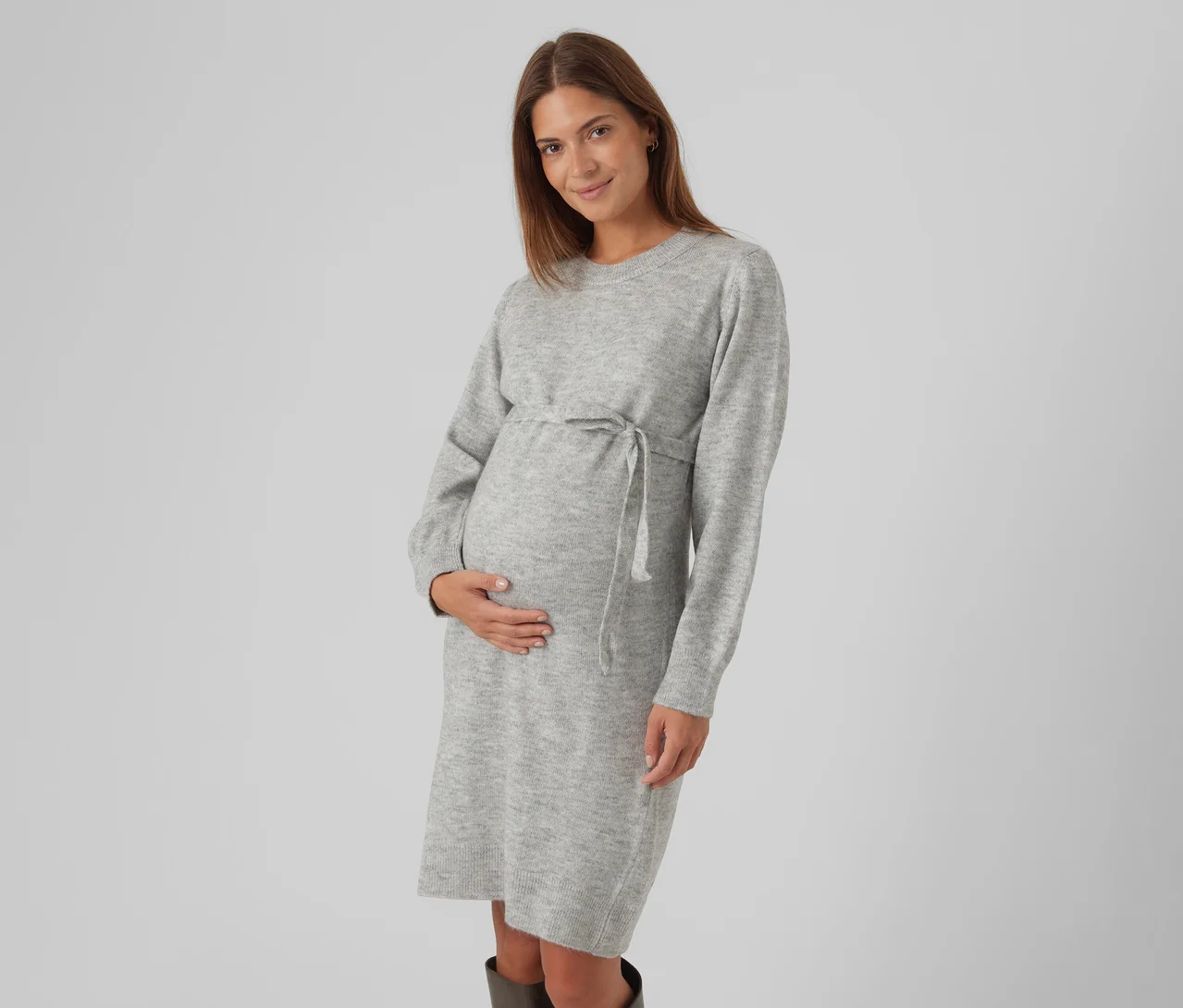 Femme enceinte aux longs cheveux bruns, portant une robe pull gris clair avec un cordon de serrage à la taille, tenant doucement son ventre et souriant.