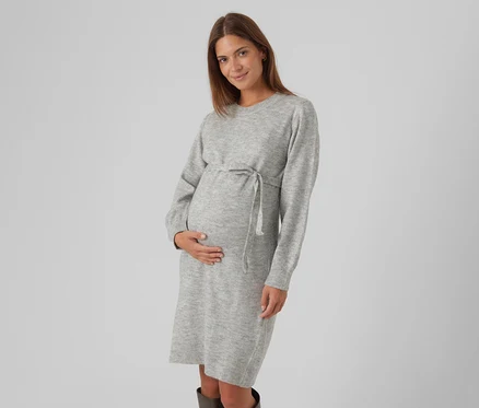 Femme enceinte aux longs cheveux bruns, portant une robe pull gris clair avec un cordon de serrage à la taille, tenant doucement son ventre et souriant.