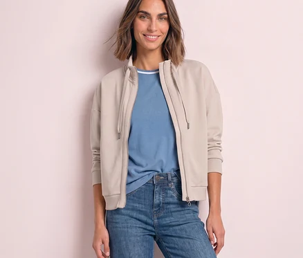 Femme souriante portant une veste de survêtement Street One beige ouverte sur un t-shirt bleu et un jean.