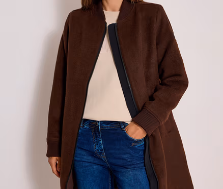 Femme souriante portant un long blouson CECIL marron, un haut beige et un jean bleu, la main dans la poche.