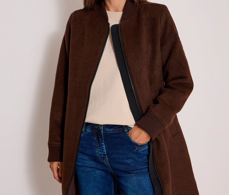 Femme souriante portant un long blouson CECIL marron, un haut beige et un jean bleu, la main dans la poche.