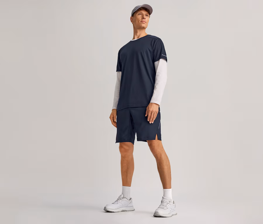Homme portant un short de sport bleu marine, un t-shirt bleu marine sur un t-shirt blanc à manches longues, des chaussettes blanches, des baskets blanches et une casquette grise.
