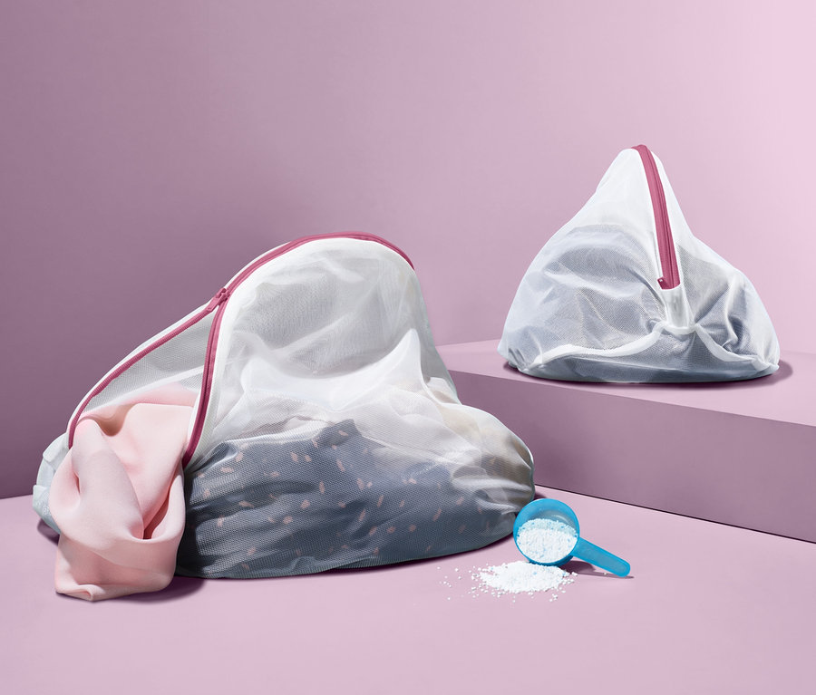 Deux filets de lavage, dont un rempli de vêtements, sont posés sur le sol à côté d'une cuillère doseuse de lessive.