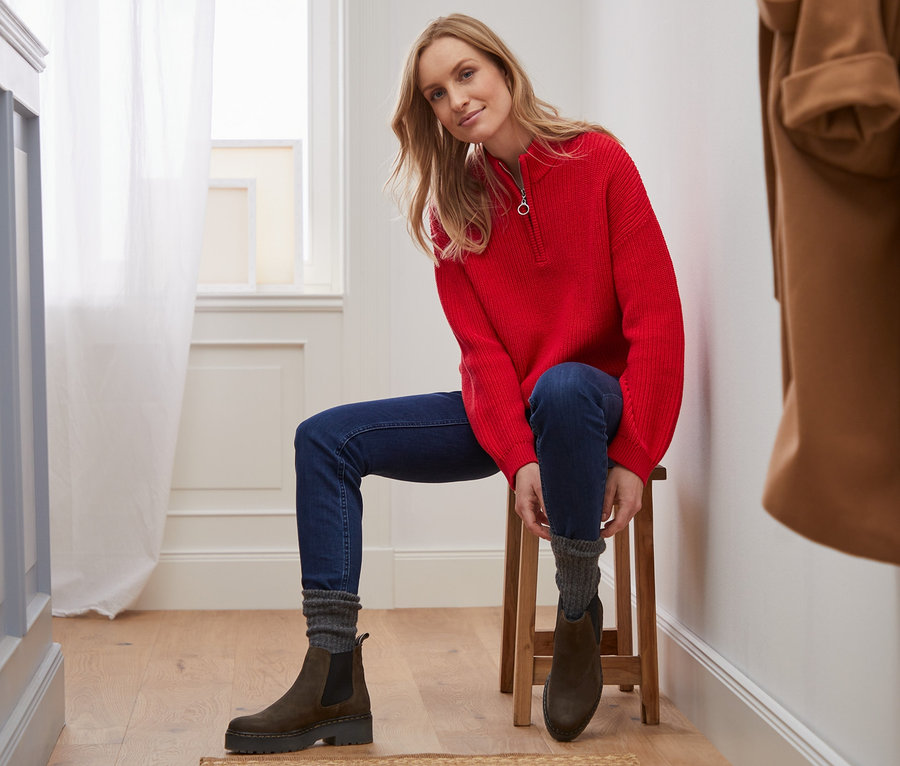 Une femme est assise sur un tabouret et porte un pull en maille avec un col, un jean et des bottes.