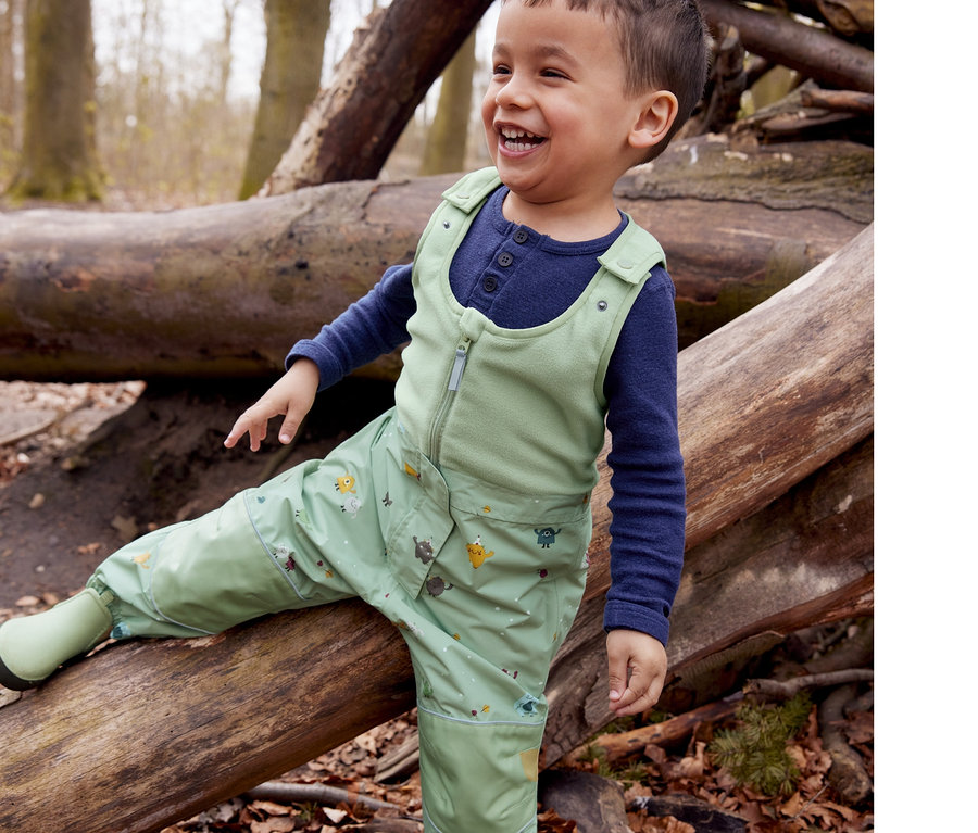 Un petit garçon souriant est assis sur un tronc d'arbre dans ses bottines de pluie Chelsea et son pantalon de pluie thermique.