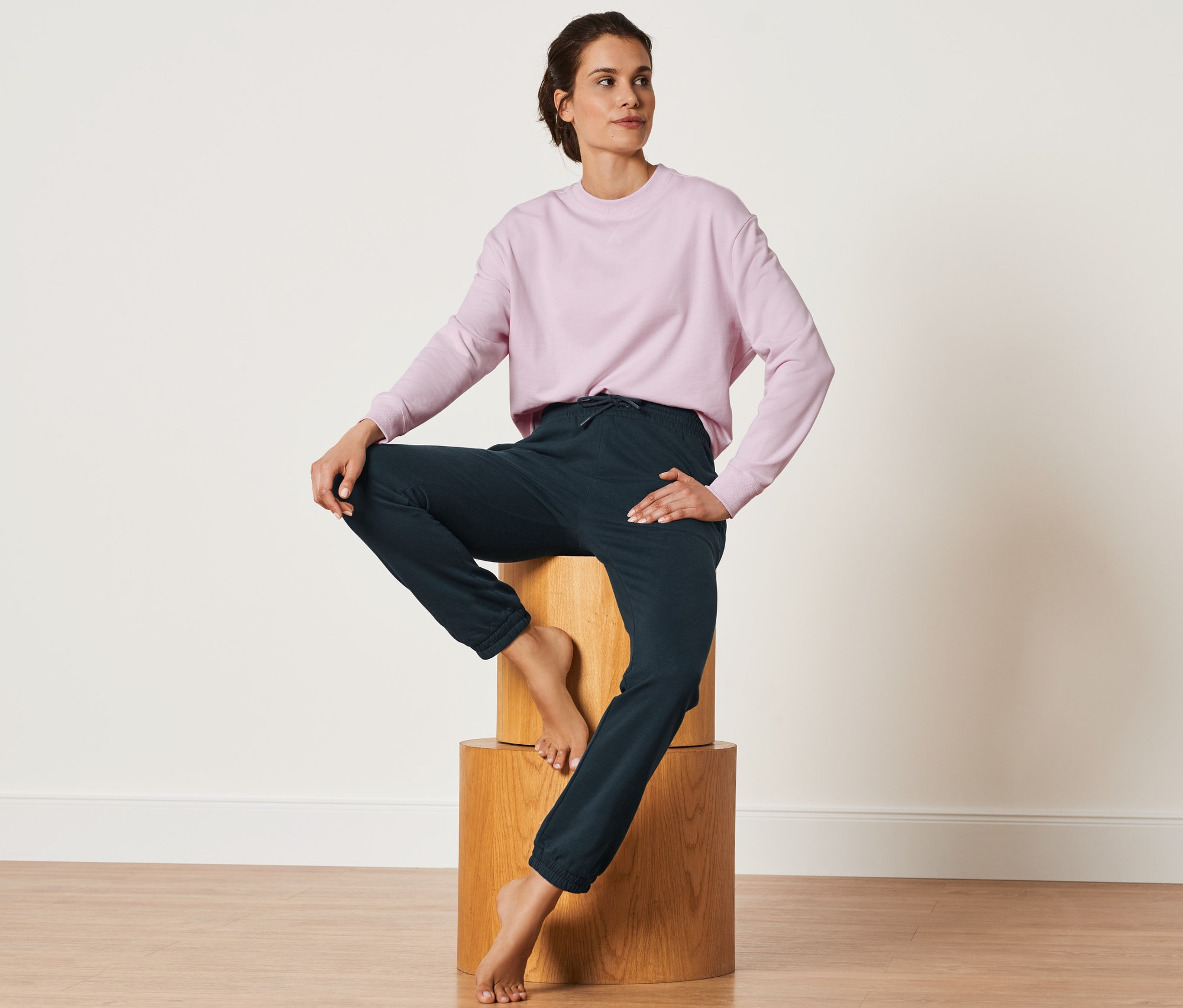 Femme assise sur des blocs de bois portant un pantalon de jogging bleu foncé et un sweat-shirt rose.