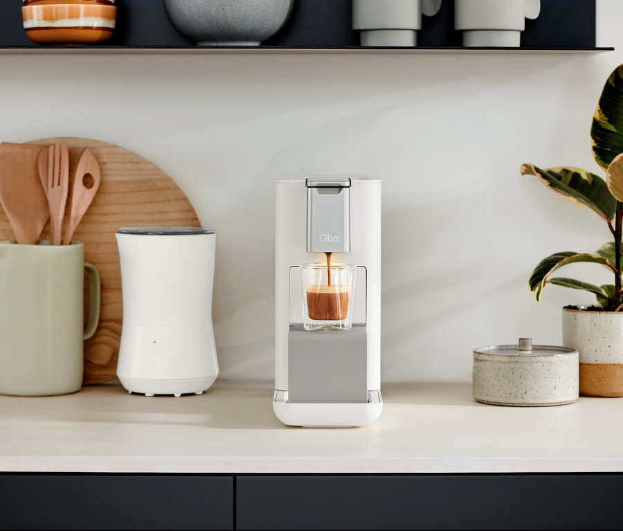 Sur un comptoir de cuisine se trouvent les machines à café Qbo ESSENTIAL Bright Sand et Qbo ICONIC Dark Chrome.