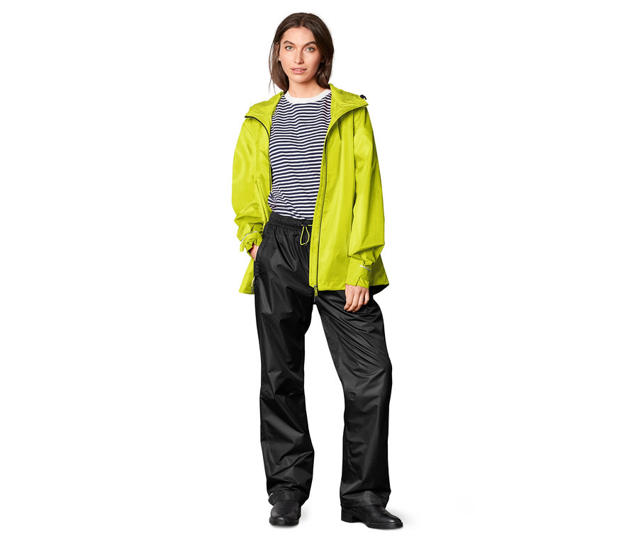 Une femme se tient sur un fond blanc et porte un pantalon de pluie noir et une veste jaune.